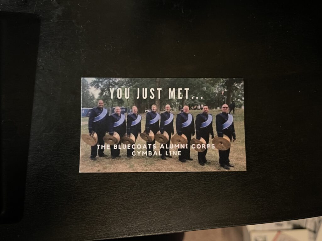 A business cards featuring a photo of the Bluecoats Alumni Corps cymbal line, in uniform and with instruments, with the caption “YOU JUST MET…THE BLUECOATS ALUMNI CORPS CYMBAL LINE”. 