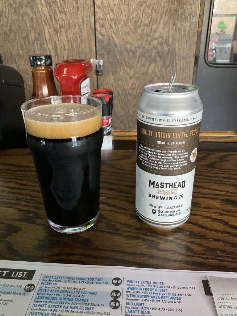 A glass of Masthead Single Origin coffee stout on the bar at the Winking Lizard. The beer is dark with a thick head on top. The beer’s can is next to the beer. 
