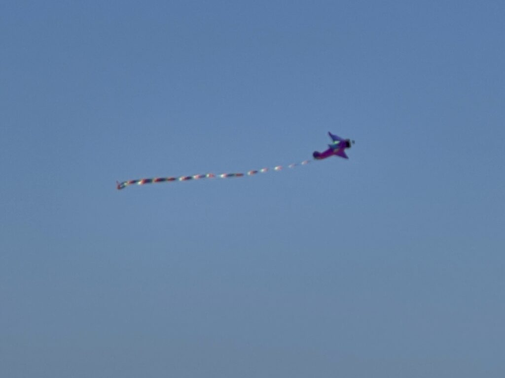 A purple kite in the shape of a biplane flying above Holden Beach. The kite has blue highlights and a long rainbow streamer trailing behind it. 