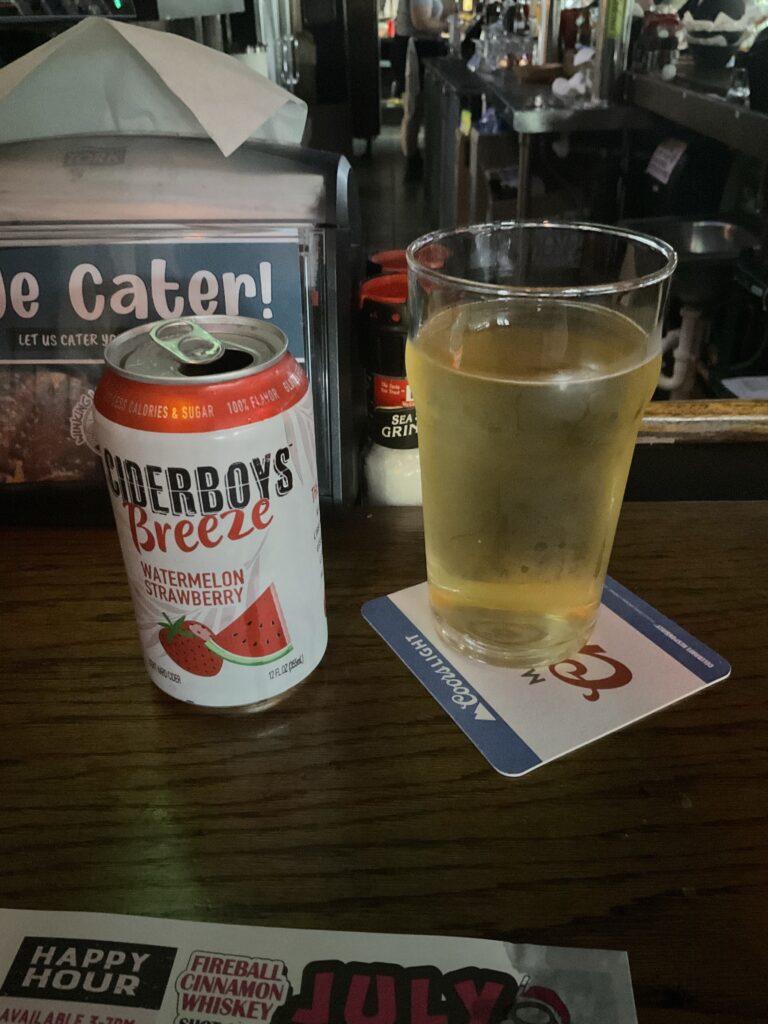 A glass of Ciderboys Breeze cider on the bar of the Winking Lizard. The cider’s can, white with a red banner along the top, features a strawberry and a watermelon slice on the front. 