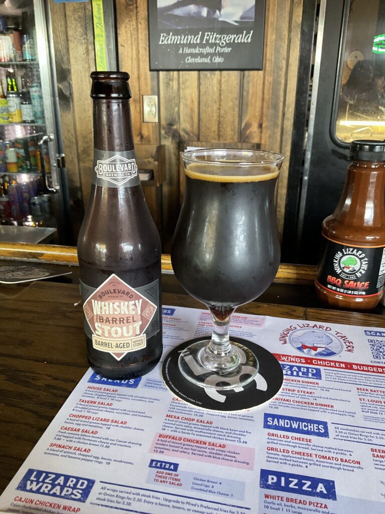 A glass of Boulevard Whiskey Barrel Stout, a dark brown beer with a hint of cream colored bubbles on top. The beer’s bottle is to the left of the glass, a similar color to the beer, with a label featuring various shades of brown and the beer’s name and logo. 