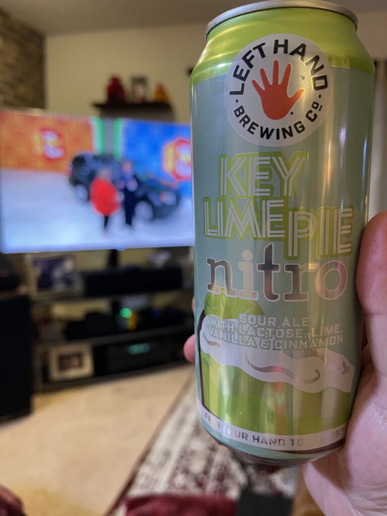 My right hand holding a can of Left Hand Key Lime Pie Nitro sour ale, with The Price Is Right on the TV in the background. 