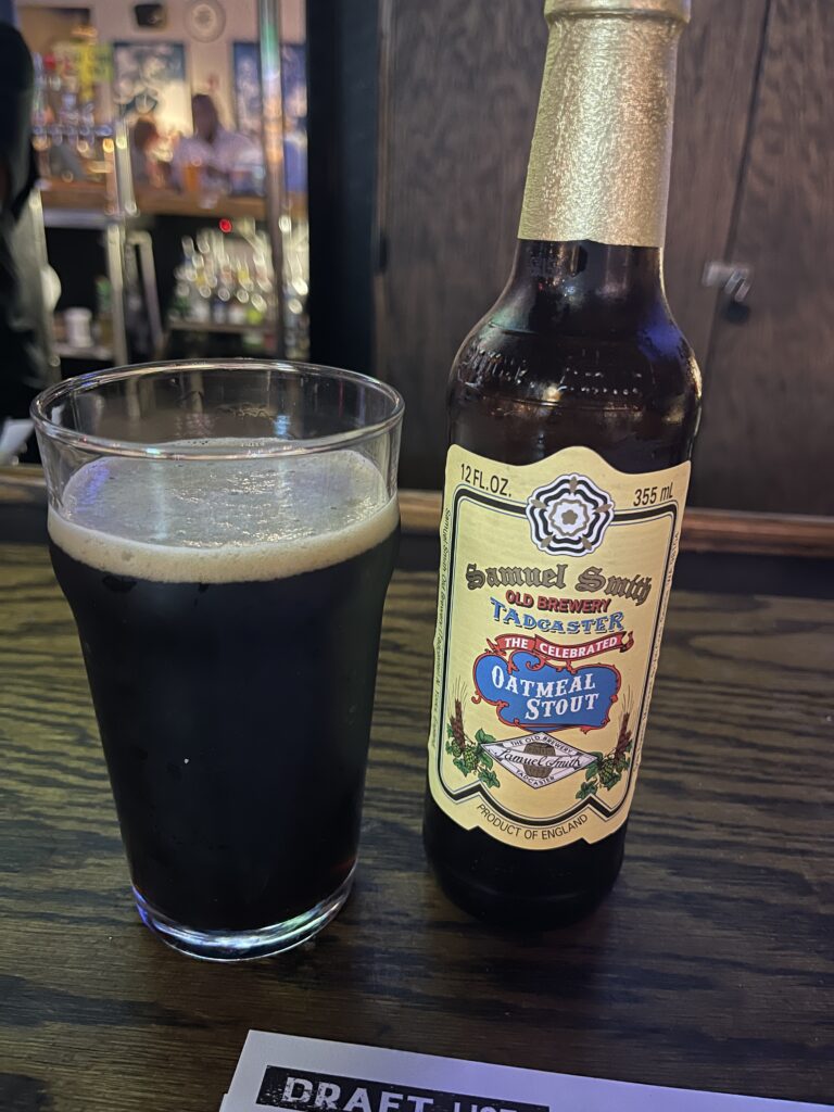 A glass of Samuel Smith Oatmeal Stout. The beer is dark brown with a small head of white bubbles on top. The beer’s bottle is to the right of the glass, with a yellow label. 