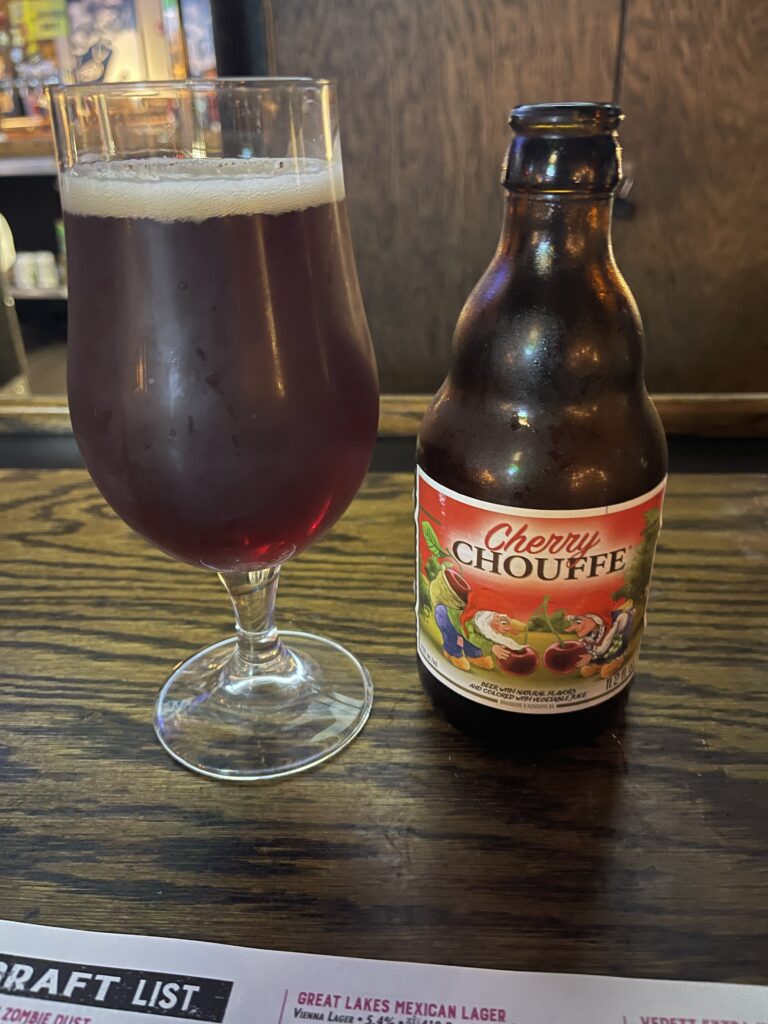 A glass of Cherry Chouffe fruit beer. This beer has a cherry red hue, topped by a small head of white bubbles. The beer’s bottle is to the right of the glass. 