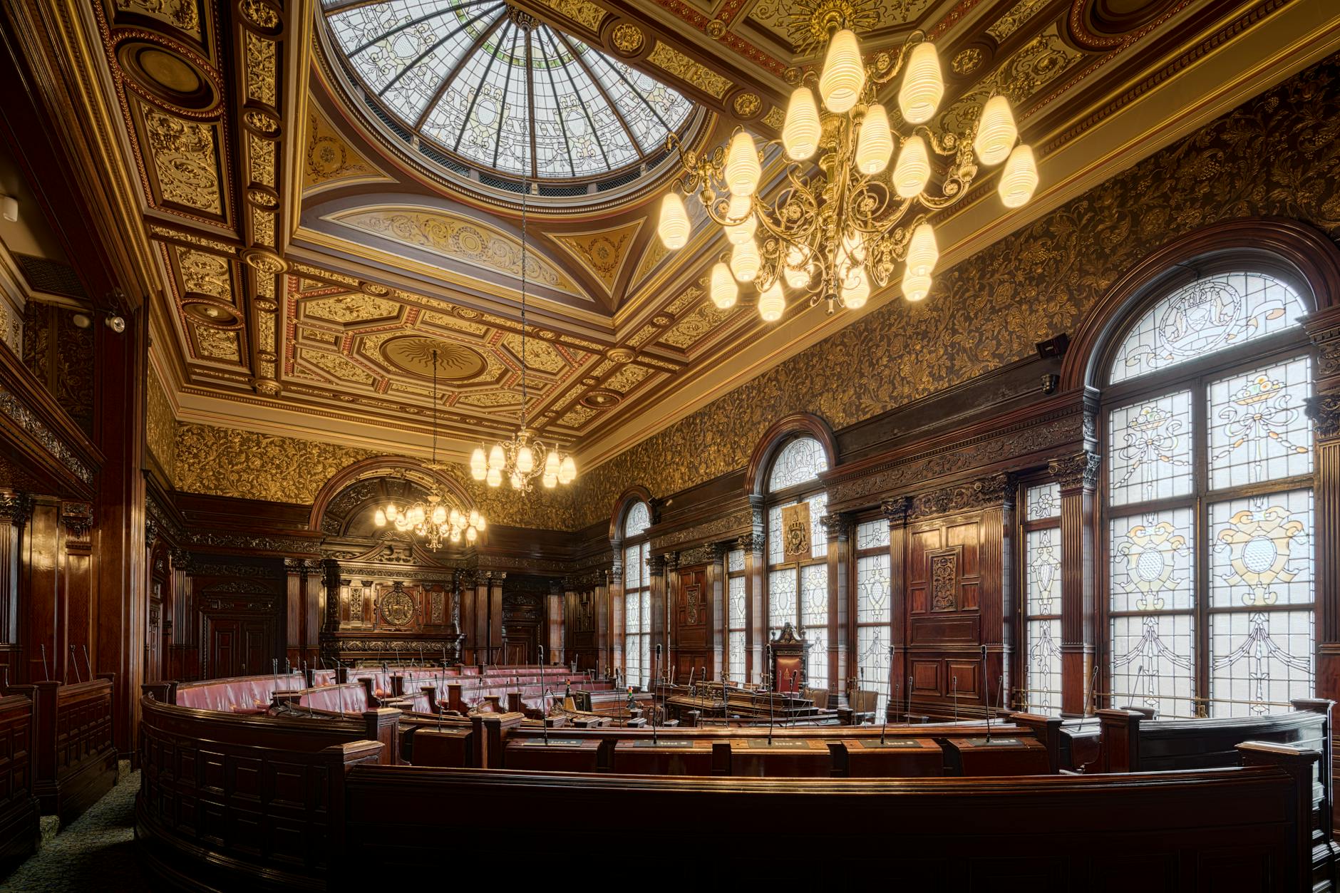 An ornate interior of a historic legislative chamber, featuring a high ceiling with a central stained glass dome, decorative walls, and wooden furnishings. The room is illuminated by vintage chandeliers and has large windows with stained glass details.